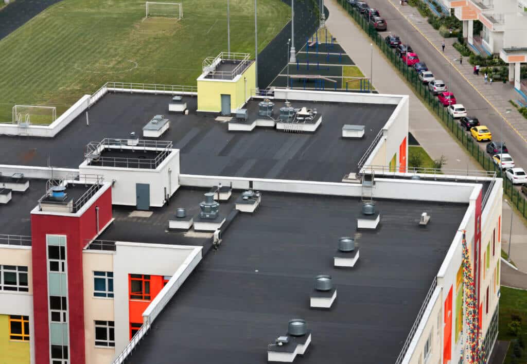 Top view flat roof with air conditioners and hydro insulation membranes on top of a modern apartment building with green football field summer day.